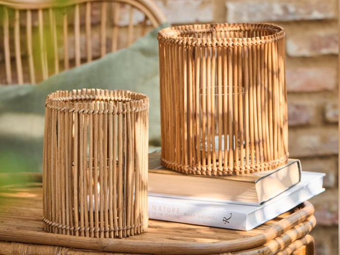 To rottinglysestager står på et rottingbord, den ene oven på to stablede bøger, med en grøn pude og en murstensvæg i baggrunden.