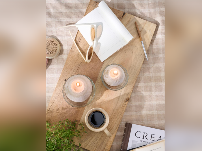 På en træbakke står to tændte stearinlys, en kop kaffe, en åben notesbog med en kuglepen, læsebriller, en plante og en delvist synlig bog med titlen CREATE.