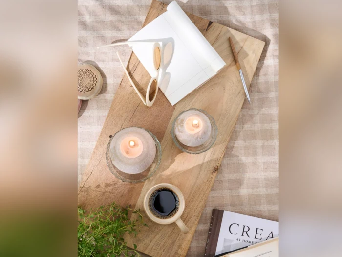 På en træbakke står to tændte stearinlys, en kop kaffe, en åben notesbog med en kuglepen, læsebriller, en plante og en delvist synlig bog med titlen CREATE.