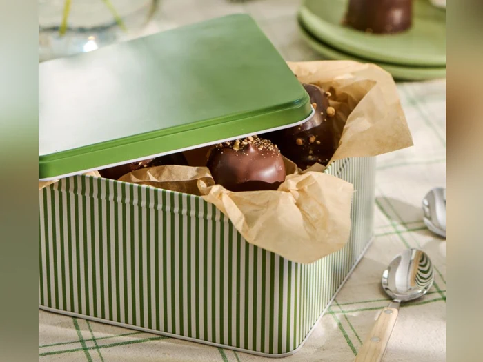 En grønstribet blikæske beklædt med brunt pergamentpapir, fyldt med chokoladeovertræk, står på et bord ved siden af to skeer.