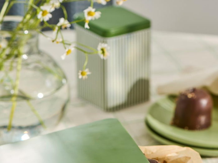 En glasvase med hvide blomster, en grøn dåse og en chokoladedessert på grønne tallerkener er arrangeret på en ternet dug.