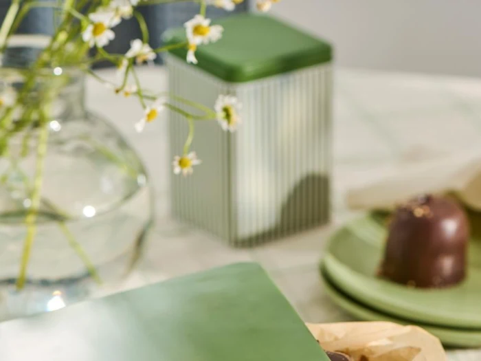 En glasvase med hvide blomster, en grøn dåse og en chokoladedessert på grønne tallerkener er arrangeret på en ternet dug.