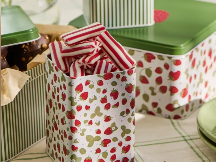 Rektangulær dåse med jordbærmønster, fyldt med rød- og hvidstribede karameller, står på et bord ved siden af andre grønne og stribede dåser.