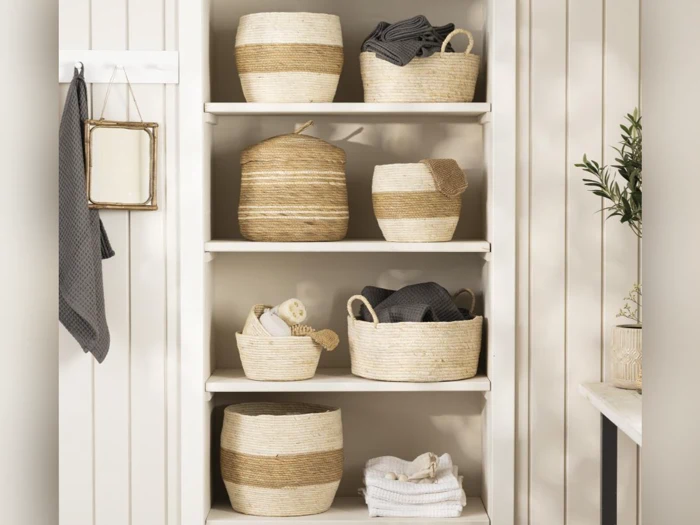 En hvid reol rummer flettede kurve, foldede håndklæder og badetilbehør. Baggrunden har lyse, lodrette paneler og en lille potteplante til højre.