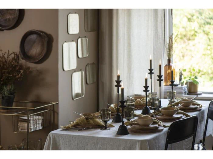 Et spisebord dækket til et måltid med stearinlys, tallerkener og glas ved siden af et vindue med lysindfald og dekorative spejle på væggen.