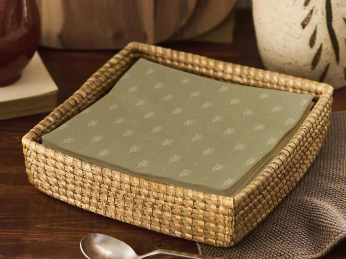 En flettet kurv rummer en stak lyse olivengrønne papirservietter med et diskret bladmønster, som er placeret på et træbord ved siden af en ske.