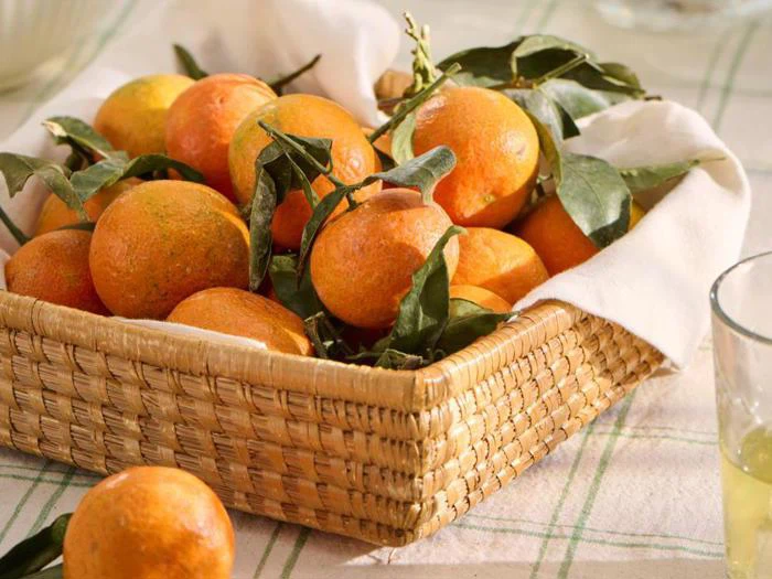 En flettet kurv fyldt med friske mandariner, nogle med grønne blade på, står på et bord med en ternet dug. Et glas juice står i nærheden.