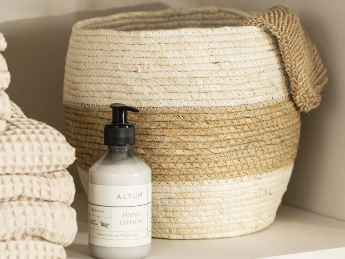 En flaske håndlotion står foran en flettet kurv med et struktureret håndklæde ved siden af på en hylde.