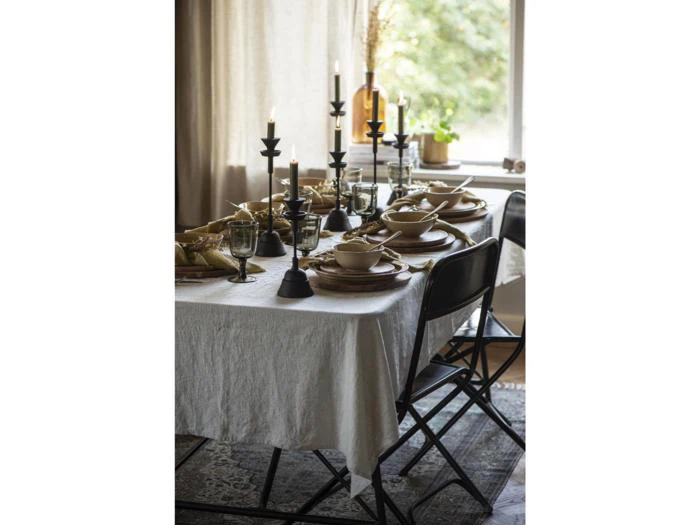 Et spisebord dækket til et måltid med beige linned, sorte stole, sorte lysestager med tændte stearinlys, tallerkener, glas og bestik, nær et vindue med naturligt lys.