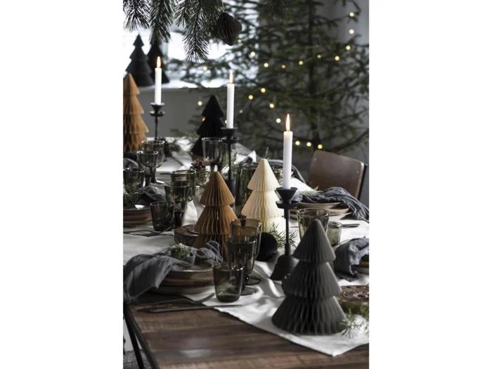 Et festligt spisebord pyntet med foldede papirjuletræer, stearinlys, glas og grønt, med et juletræ og lyskæder i baggrunden.