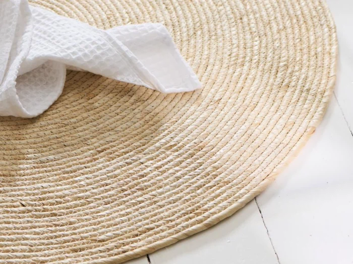 Et rundt, vævet stråtæppe på et hvidt trægulv med et hvidt, struktureret klæde delvist oven på.