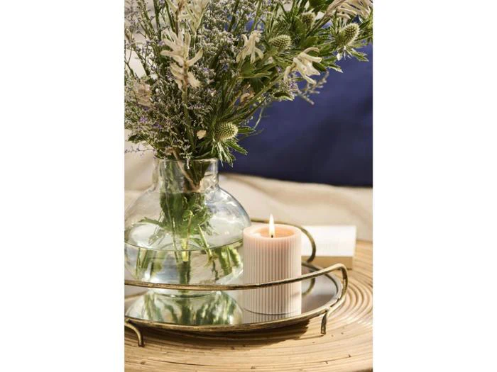 En glasvase med hvide blomster og grønt står på en rund spejlbakke ved siden af et tændt hvidt ribbelys.