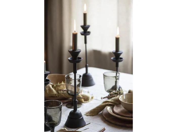 Tre sorte lysestager med tændte stearinlys er anbragt på et dækket spisebord med glas, tallerkener og servietter i et blødt oplyst rum.