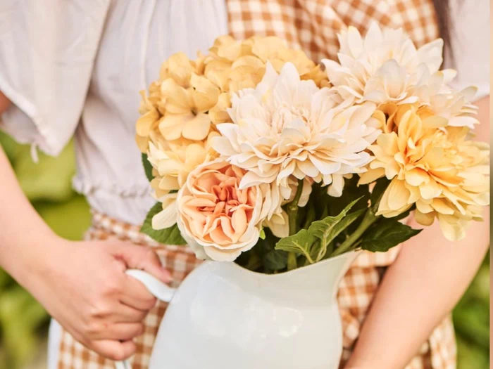 En person i gingham-kjole holder en hvid kande fyldt med cremefarvede og gule blomster, bl.a. georginer og roser.