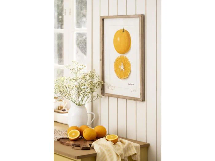 Et køkkenbord med hele og halve appelsiner, en hvid kande med blomster og en indrammet illustration af appelsinfrugter, der hænger på en væg med hvide paneler.
