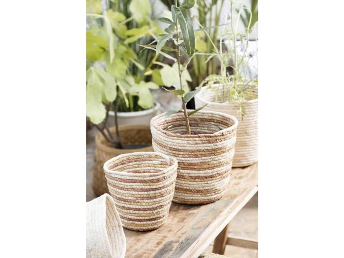 To flettede kurve med diskrete rosa og beige striber er placeret på en træoverflade, og i den ene står en potteplante. Andre potteplanter og grønne planter er synlige i baggrunden.