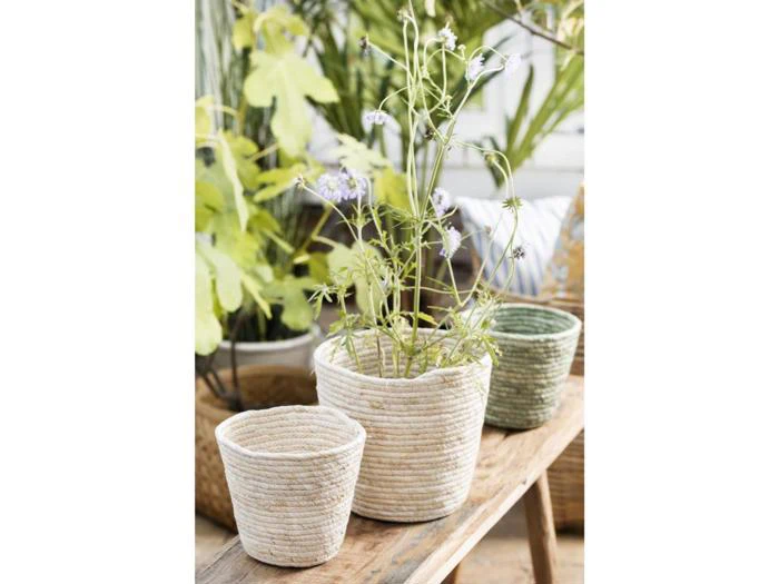 Tre flettede kurve står på en træbænk; den ene indeholder en blomstrende plante med sarte lilla blomster, mens de to andre er tomme. I baggrunden ses frodigt grønt løv.