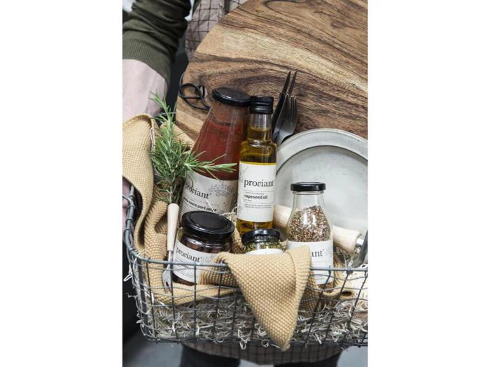 En trådkurv med et glas sauce, olivenolie, krydderier, et skærebræt, en tallerken, redskaber, et glas marmelade, en rosmarinkvist og et solbrunt klæde.