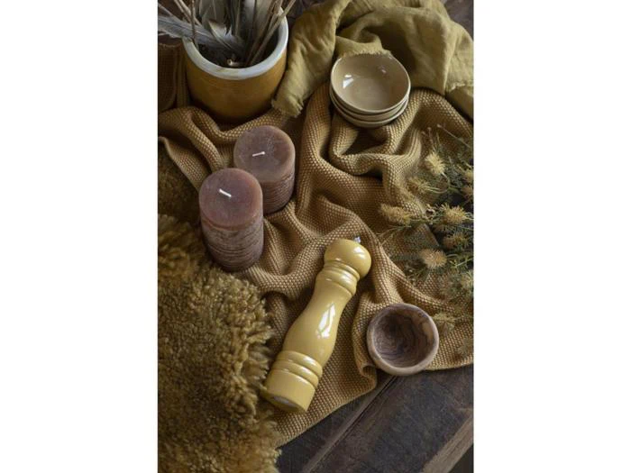 En gul peberkværn, to brune stearinlys, beige skåle, en struktureret sennepsklud, tørrede blomster og et loddent tæppe på en træoverflade.