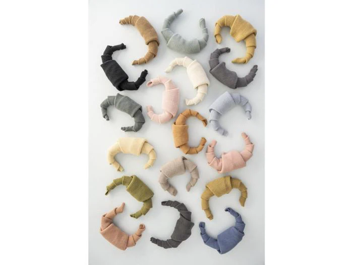 Udstoppet stoflegetøj formet som croissanter i forskellige pastelfarver og neutrale farver, arrangeret i pæne rækker på en hvid baggrund.