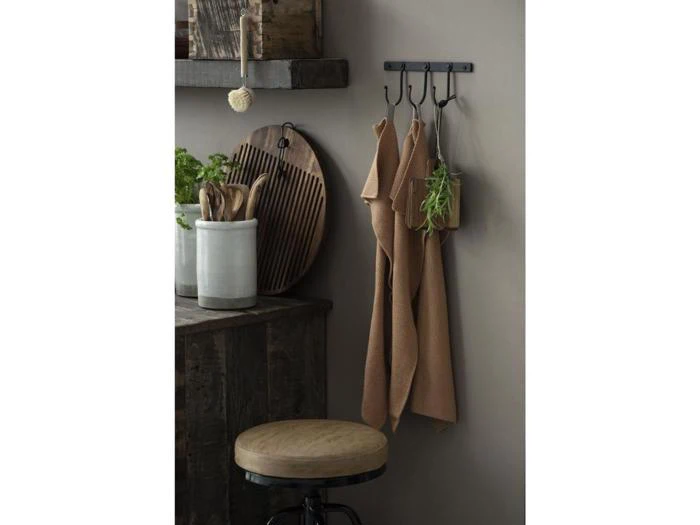 En rustik køkkenscene med en træskammel, redskaber i en keramikkrukke, skærebrætter og brune håndklæder, der hænger på kroge; der hænger også krydderurter på en krog.