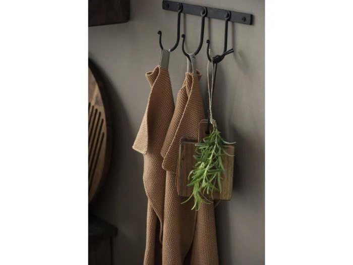 To brune håndklæder med struktur hænger på sorte metalkroge ved siden af et lille skærebræt i træ med en grøn kunstig plante på en beige væg.