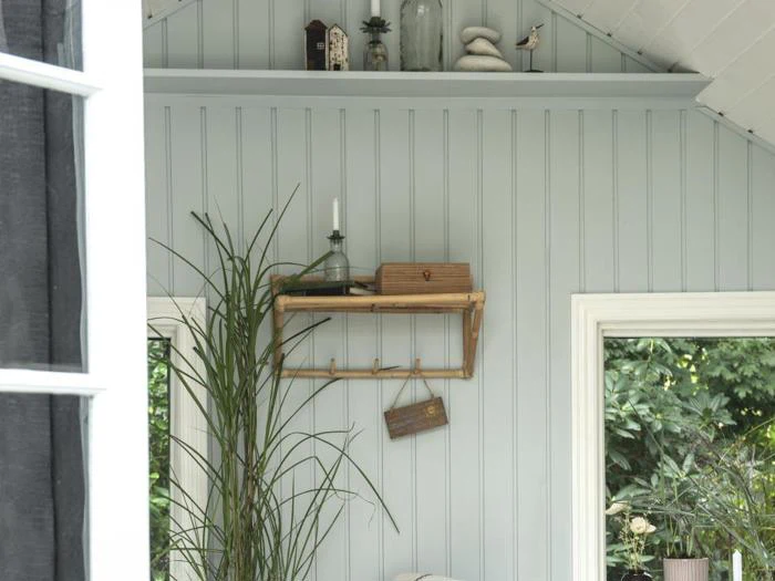 En væg med lyseblå paneler og en træhylde med et stearinlys, en glasflaske og en lille æske. En høj grøn plante og et stort vindue med grønt udenfor er synlige.