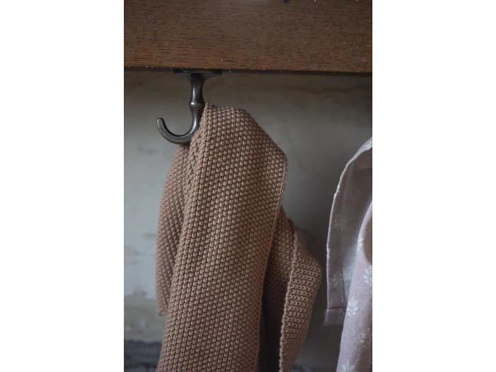 Et brunt håndklæde med struktur hænger på en metalkrog på en træoverflade, og en del af et lyst stof er synligt til højre.