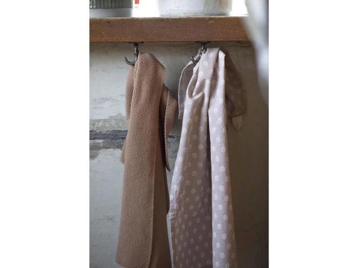 To mønstrede beige klude hænger side om side på sorte kroge mod en hvid trævæg.