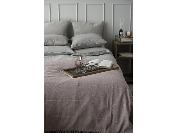 En pænt redt seng med grå puder, et lilla tæppe og en træbakke med glas, et stearinlys og en åben bog. Ved siden af sengen står et natbord med stearinlys og dekorationer.