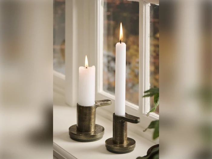 To tændte hvide lys i messingholdere står i en vindueskarm med blødt dagslys ind gennem vinduet og en grøn plante delvist synlig til højre.