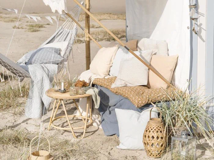Hyggelig opsætning på stranden med en polstret bænk, et flettet bord, en lanterne og dekorative puder og tæpper under et telt, omgivet af sand og græs.