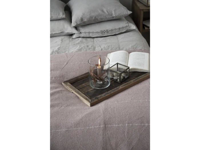 På en træbakke på en seng står et tændt stearinlys i en glasholder, en åben bog og en lille æske med æg. Sengen har gråt sengetøj og flere puder.