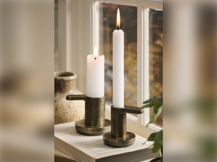 To tændte hvide stearinlys i metalholdere står på en åben bog ved et vindue med en keramikvase og en grøn plante i nærheden. Varmt lys filtreres gennem glasset.