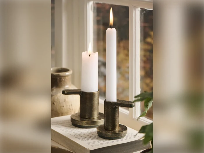 To tændte hvide stearinlys i metalholdere står på en åben bog ved et vindue med en keramikvase og en grøn plante i nærheden. Varmt lys filtreres gennem glasset.