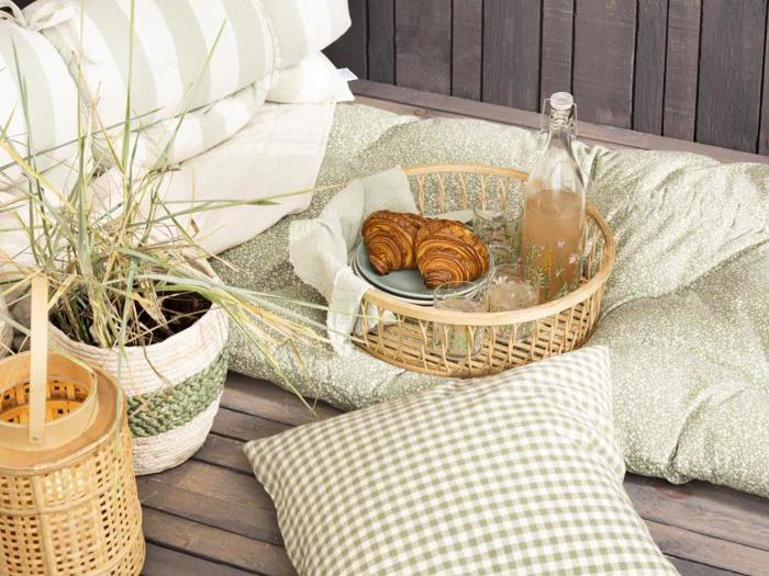 Et hyggeligt udendørs setup med hynder, en plante og en flettet bakke med croissanter, glas og en flaske juice på et trægulv.