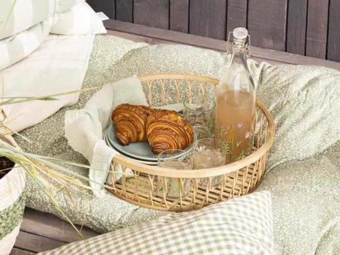 En flettet bakke med croissanter, to glas og en flaske juice på en grøn pude med ternede og blomstrede puder ved siden af.