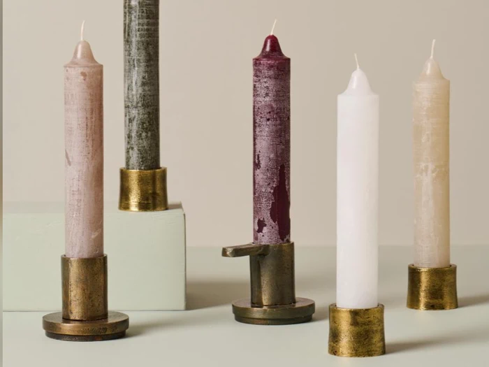 Fem høje, strukturerede lys i forskellige farver med guldlysestager, arrangeret på en lys flade mod en neutral baggrund.