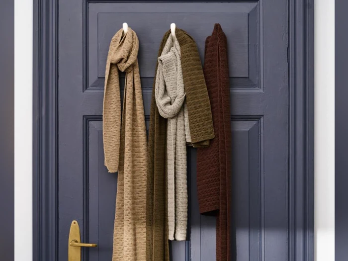 Tre strukturerede tørklæder i beige, olivengrøn og brun hænger på hvide kroge på en mørkeblå dør med messinghåndtag.