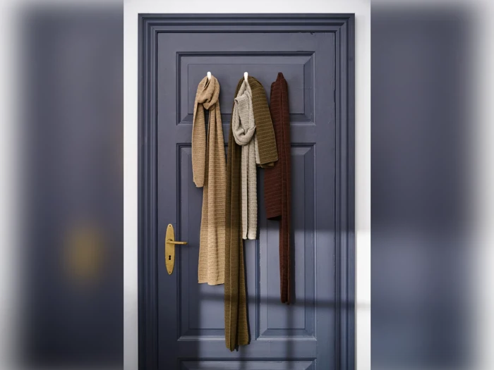 Tre strikkede tørklæder i brune og beige nuancer hænger på kroge på en lukket mørkeblå dør med guldhåndtag, oplyst af blødt naturligt lys.
