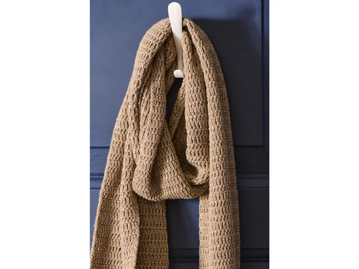 Et struktureret beige strikket tørklæde hænger på en hvid krog mod en mørkeblå væg.