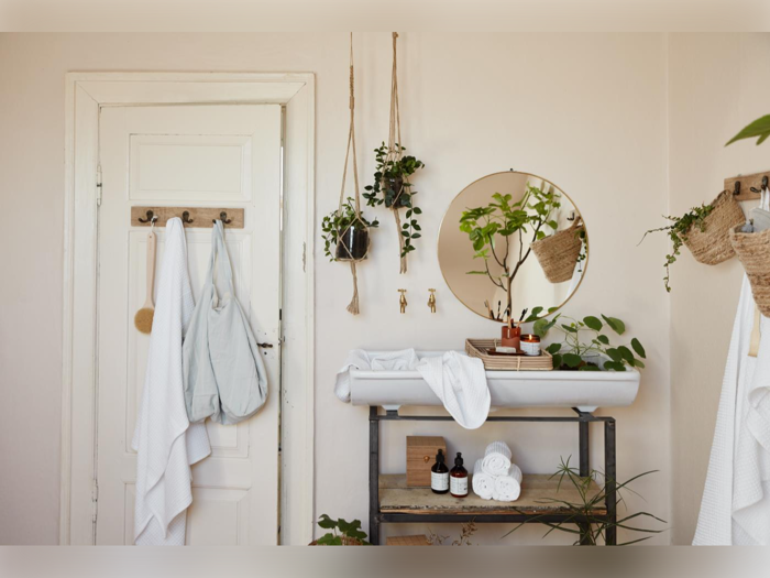 Et hyggeligt badeværelse med hængende planter, et rundt spejl, håndklæder på stativ og kroge, hylder med toiletartikler og en hvid håndvask under spejlet.