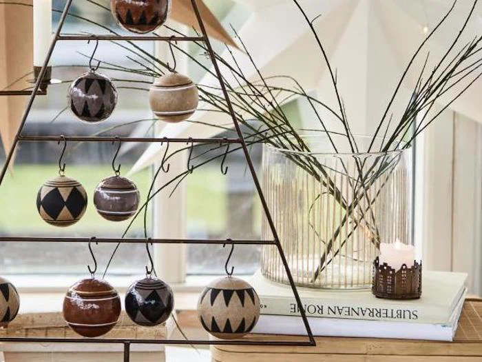 Et trekantet metalstativ viser mønstrede ornamenter ved siden af en glasvase med kviste og et tændt stearinlys på stablede bøger nær et vindue.