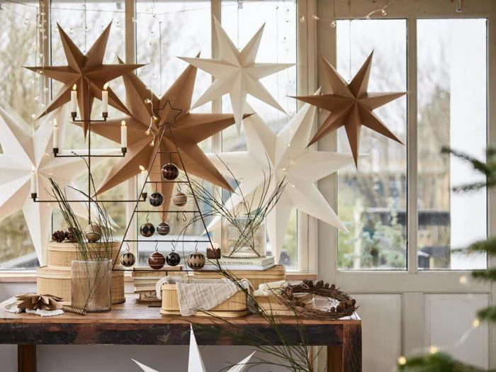 Et festligt bord ved et vindue pyntet med hvide og brune papirstjerner, stearinlys, pynt, kogler og naturgrene.