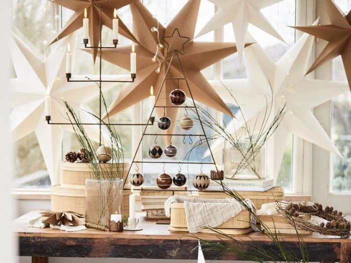 Et minimalistisk juletræ i ståltråd med brune og hvide ornamenter står på et træbord omgivet af stearinlys, kogler og store papirstjerner ved et vindue.