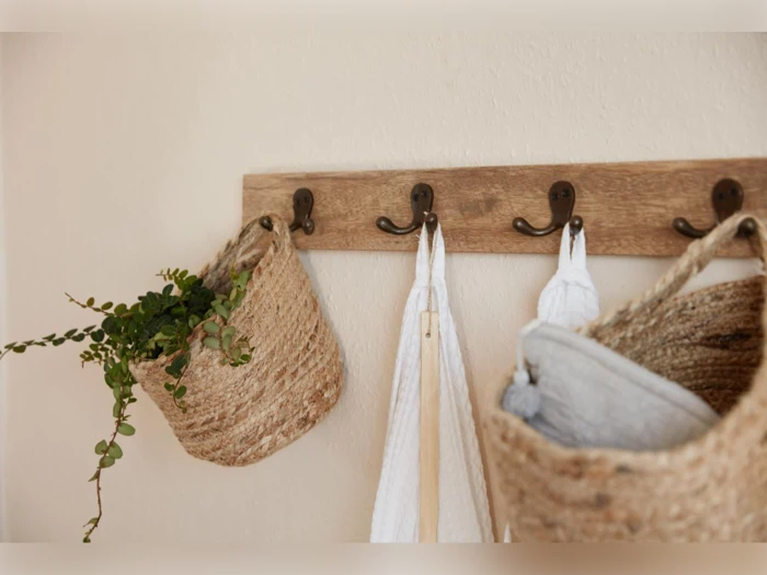 Et vægstativ i træ med metalkroge holder flettede kurve, en plante og hvide håndklæder op mod en beige væg.