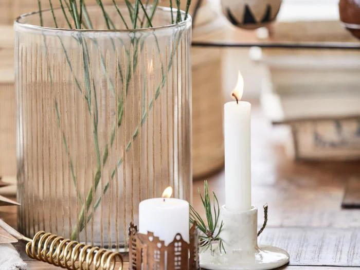 Et tændt hvidt lys i en holder, et mindre tændt lys med et dekorativt hylster og en glasvase med grønne stilke på en træplade.