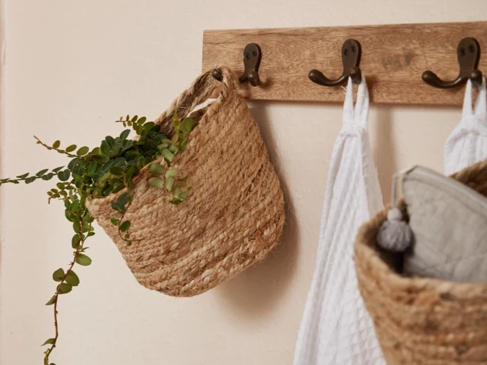 Et vægstativ af træ med tre kroge holder en flettet kurv med grønne blade, et hvidt håndklæde og en anden flettet kurv med gråt stof.