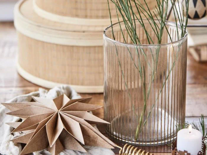 Klar ribbet glasvase med tynde grønne stilke, en foldet brun papirstjerne, et tændt hvidt lys og en rund kurvebeholder på en træoverflade.