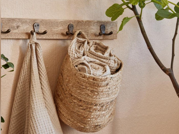 Et beige håndklæde og en flettet kurv med sammenrullede håndklæder hænger på vægkroge ved siden af en grøn plante.